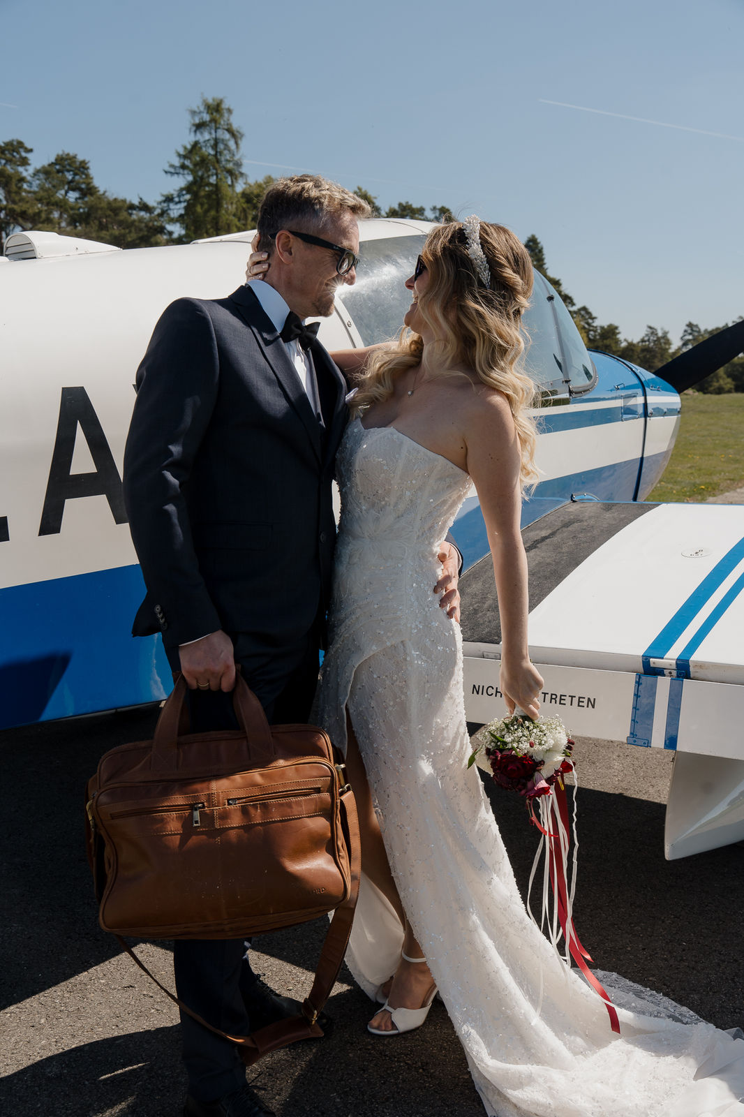 Brautpaar mit Reisetasche vor einem kleinen Flugzeug am Flugplatz
