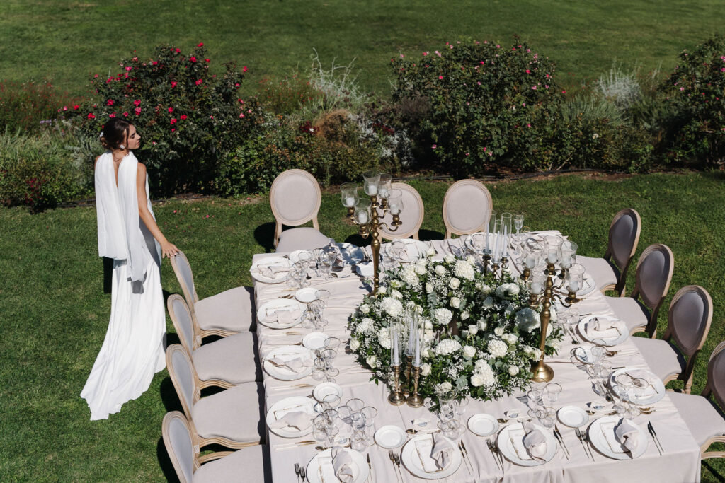 Braut neben der rechteckigen Hochzeitstafel mit weißer Floristik im Garten der Vigna Caio