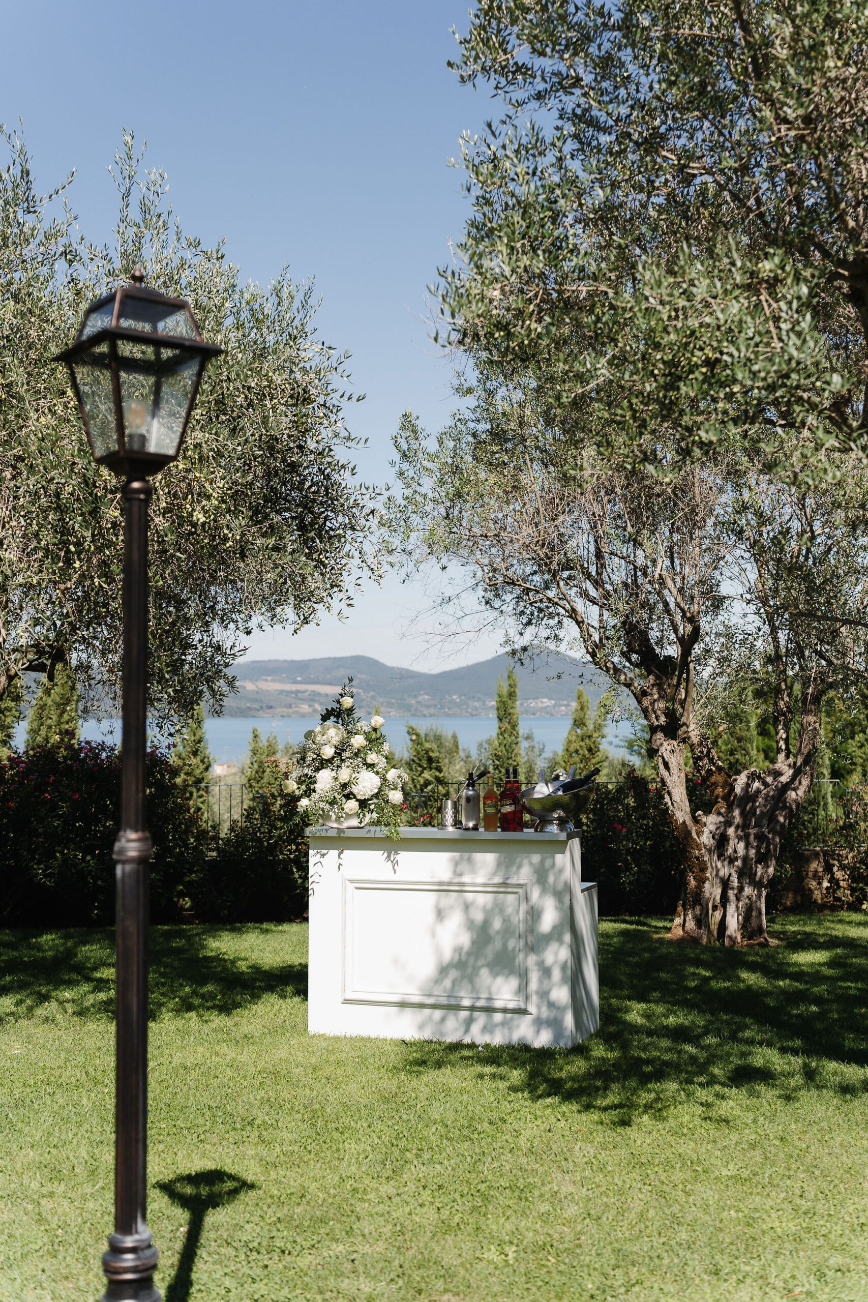 Weiße Hochzeitsbar mit Blumen und Getränken zwischen Olivenbäumen mit Blick auf den Braccianosee