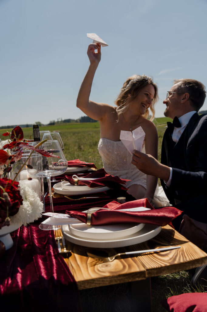 Braut und Bräutigam sitzen lachend an einem rot dekorierten Hochzeitstisch im Freien
