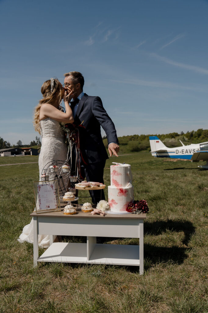 Brautpaar neben der Hochzeitstorte auf einer Wiese am Flugplatz